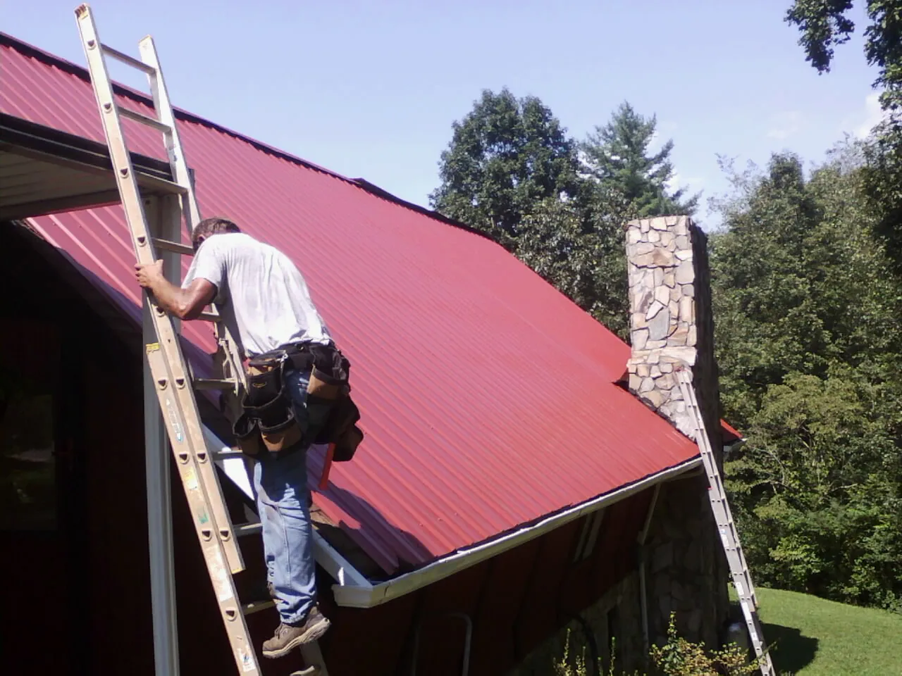 Roofer on a ladder beside a red metal roof and stone chimney — Soffit & Fascia Repair by Roofing Brothers in Babson Park